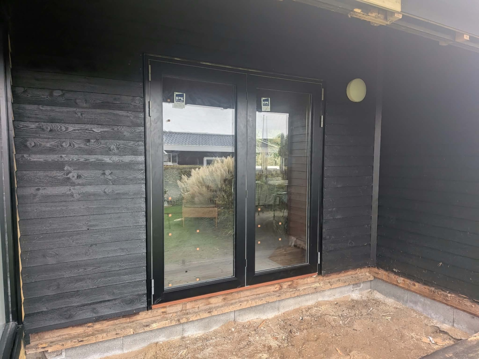 Dobbeltdør i glas åbner til have, grå træfacade og indvendigt gulv under opbygning.
